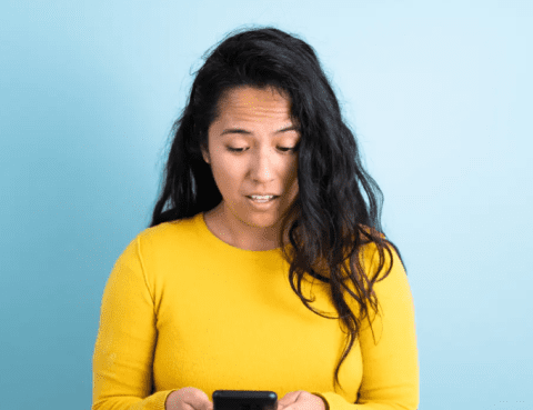 woman with long dark hair looking at her phone in shock