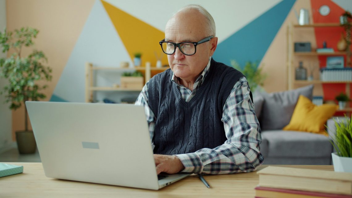 A man with thinning white hair and black-framed glasses sits at a desk while working on a laptop.