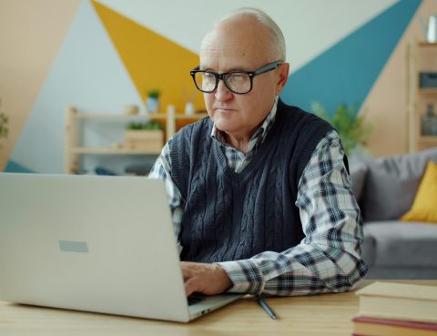 A man with thinning white hair and black-framed glasses sits at a desk while working on a laptop.