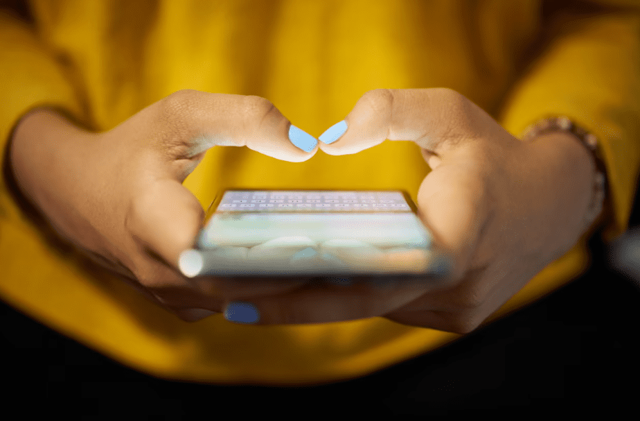 A close up of the hands of a woman wearing a yello as she holds her cell phone