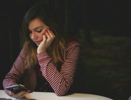 A woman leaning with her elbows on a table looking down at a cellphone with a worried expression, in a dark room