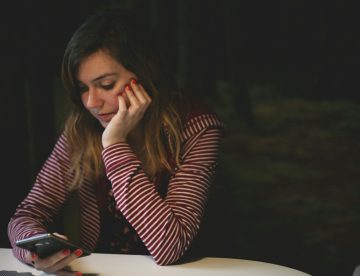 A woman leaning with her elbows on a table looking down at a cellphone with a worried expression, in a dark room