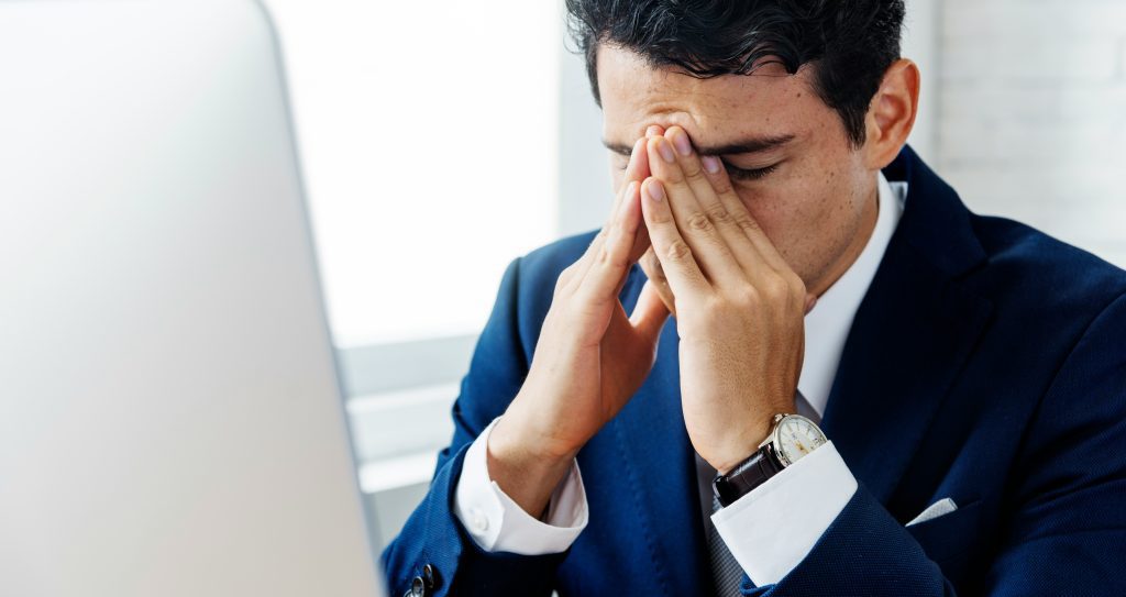 man in business suit sit's at his desk looking stressed with his hands over his face