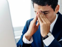 man in business suit sit's at his desk looking stressed with his hands over his face