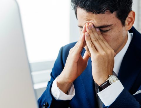 man in business suit sit's at his desk looking stressed with his hands over his face