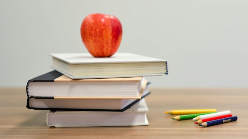 a stack of books with a red apple on the top beside a few colored pencils