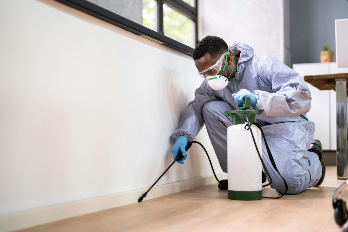 A pest control worker in protective clothing and a mask sprays pesticide onto the baseboard of a house.