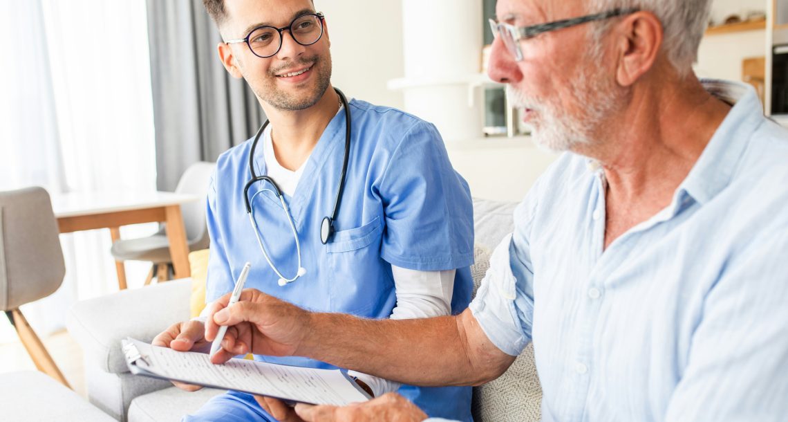 A senior man sits next to a nurse who wears blue scrubs as they talk about paperwork on a clipboard