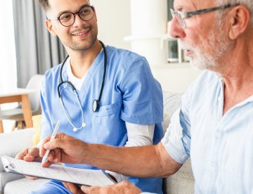 A senior man sits next to a nurse who wears blue scrubs as they talk about paperwork on a clipboard
