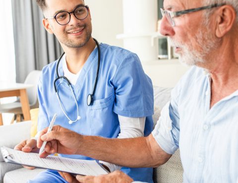 A senior man sits next to a nurse who wears blue scrubs as they talk about paperwork on a clipboard