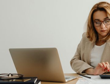 woman using a calculator at her desk which has a laptop and paperwork on it