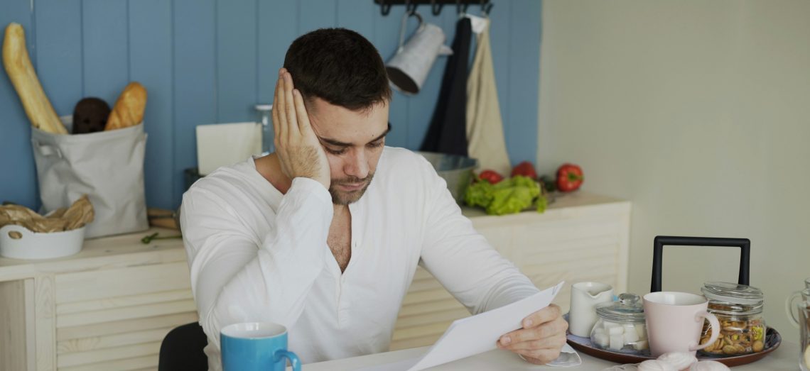 A man at a breakfast table looks at a letter with worry on his face