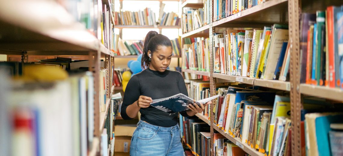 A young woman reads from a book while standing surrounded by packed book shelves