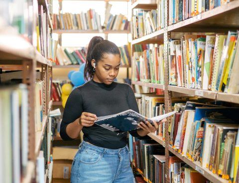 A young woman reads from a book while standing surrounded by packed book shelves