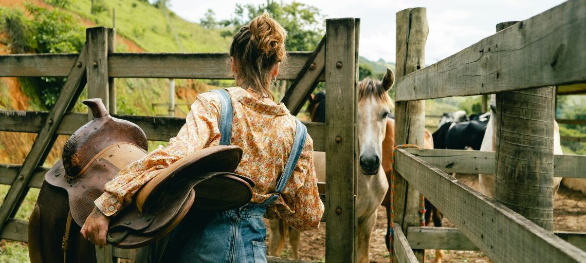 A woman holding a saddle opens a fence gate where a horse waits for her