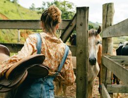 A woman holding a saddle opens a fence gate where a horse waits for her