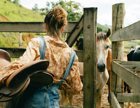 A woman holding a saddle opens a fence gate where a horse waits for her
